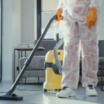 A cleaner in protective coveralls and gloves vacuuming a modern living room.