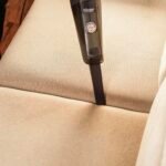 A person using a handheld vacuum to clean a beige sofa indoors, showcasing effective cleaning.