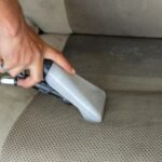 Hands using a vacuum nozzle to deeply clean a fabric car seat, removing dirt and stains.